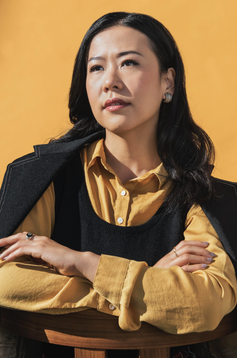 Domee Shi arms crossed and resting on back of a chair looking pensive against a yellow backdrop